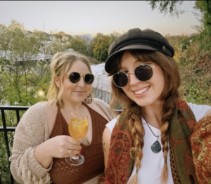 Two women smiling outdoors. One holds a drink, both wear sunglasses. The setting is a scenic, wooded area with soft evening light, conveying a relaxed, joyful mood.