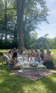 A group of people sits on blankets having a picnic in a sunny park. They are gathered around a cloth-covered table with food and drinks, under a large tree.